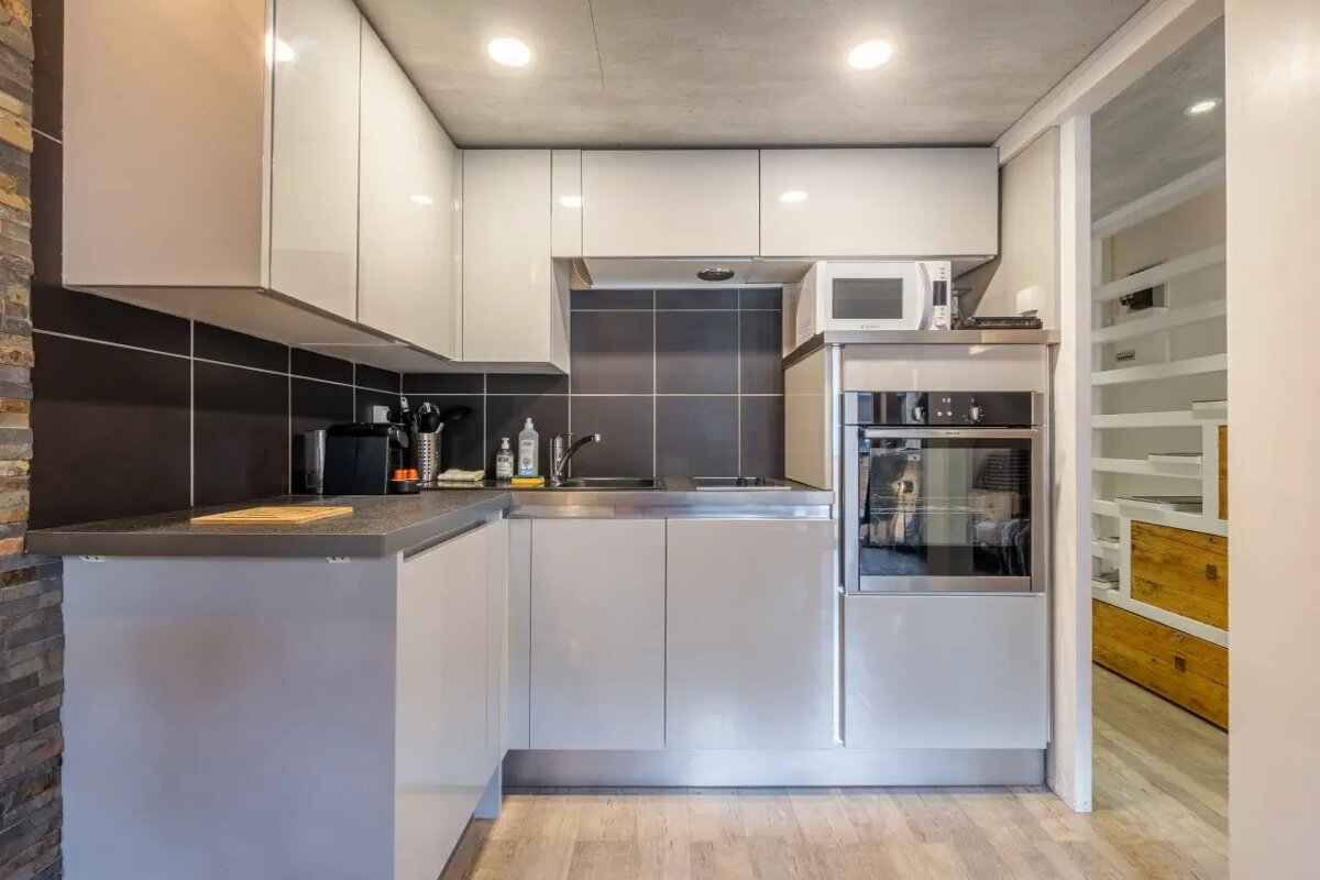 Compact modern kitchen with sleek light gray cabinets, a black tiled backsplash, built-in appliances, and light wood flooring.