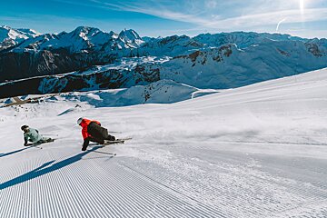 Two skiers carve on a groomed snowy slope, spraying snow. Majestic mountains tower under a clear blue sky.