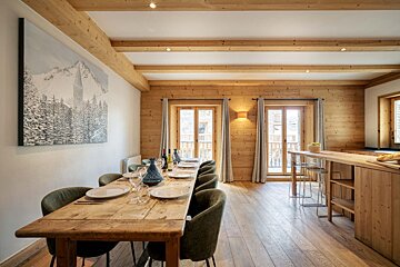 A cozy chalet dining room with a long wooden table, green chairs, exposed ceiling beams, and a large snowy mountain painting on the wall.