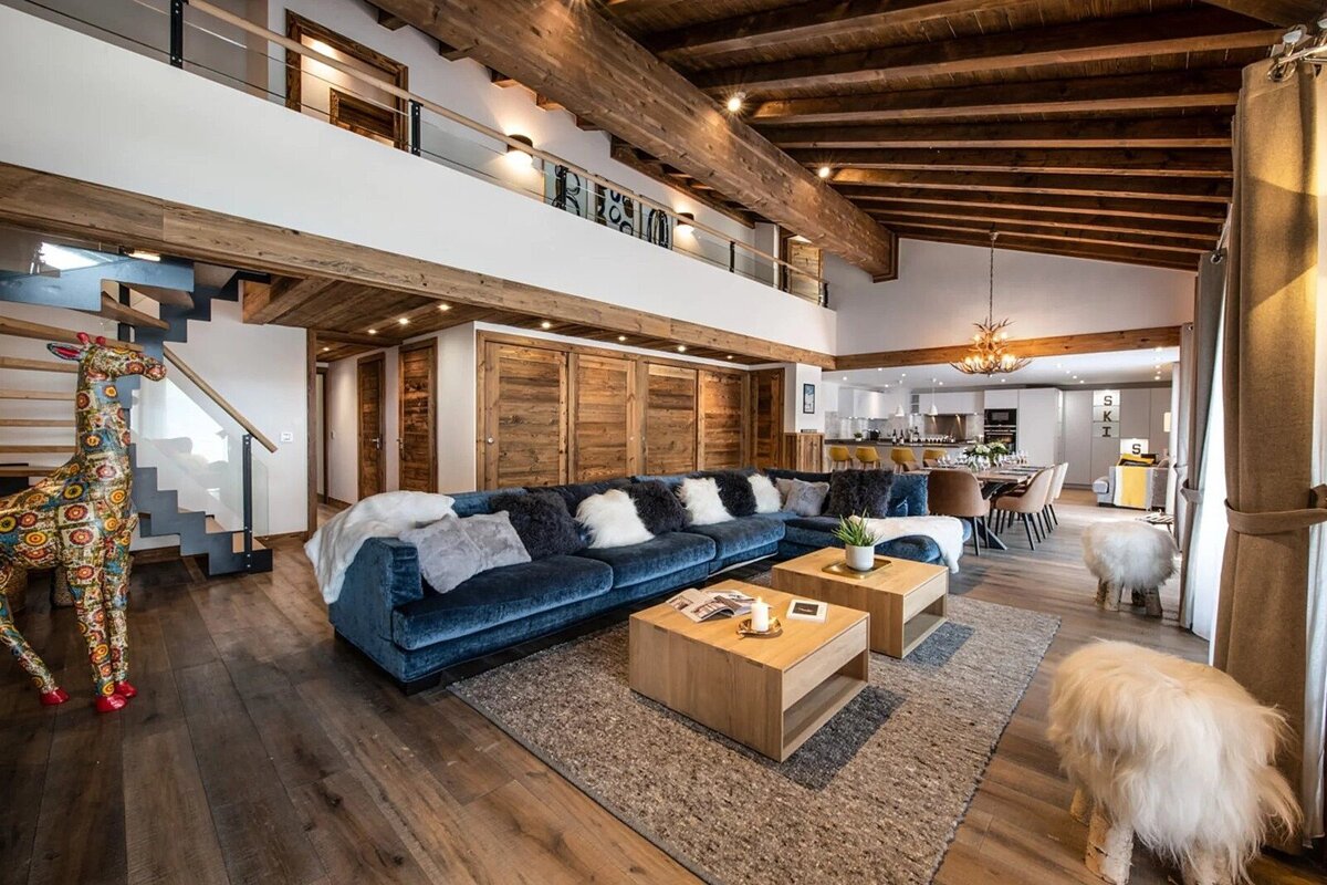 Luxurious chalet-style living room with a large blue sectional, wooden beams, loft, open kitchen/dining, and a whimsical giraffe statue.
