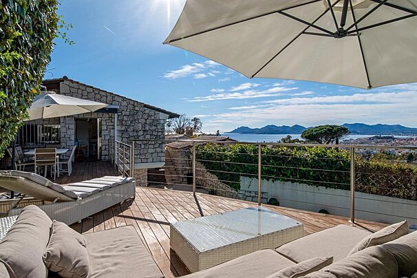 A sunny wooden terrace with lounge chairs, umbrellas, and a glass-top table overlooks a coastal view under a bright blue sky.