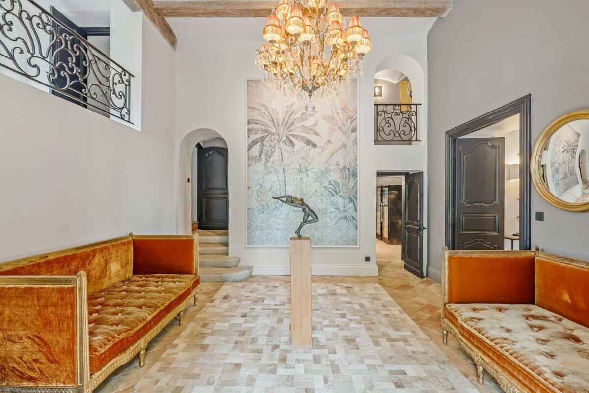 A grand, high-ceilinged entryway with a lavish chandelier, tropical mural, sculpture, and two ornate orange velvet sofas. Arched doorways and balconies create a luxurious feel.