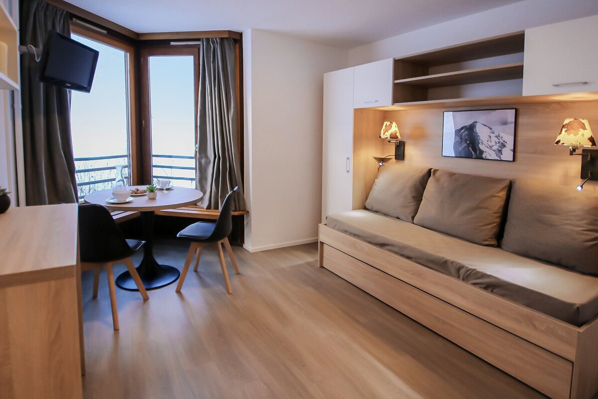 A bright, modern studio apartment featuring a dining area by a scenic window and a comfortable daybed with built-in lighting and storage.