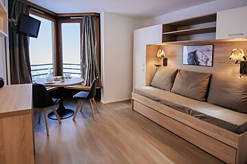 A bright, modern studio apartment featuring a dining area by a scenic window and a comfortable daybed with built-in lighting and storage.