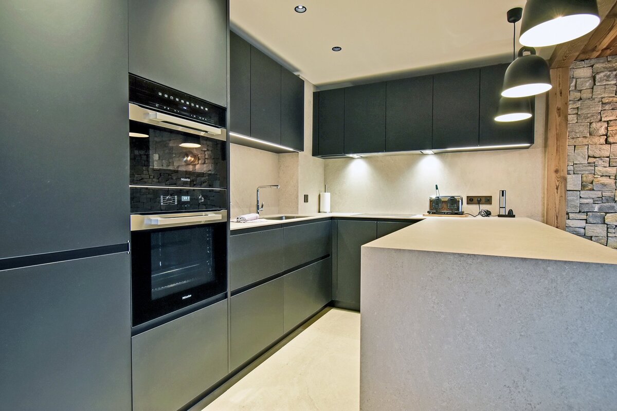 Modern kitchen with dark grey cabinets, built-in ovens, light grey countertops/island, and a stone wall. Under-cabinet & pendant lights illuminate the space.