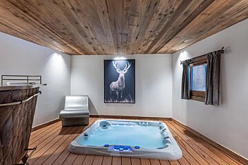 An indoor hot tub room with wooden floors and ceiling, a silver lounge chair, a bar, and a large stag painting on the wall.