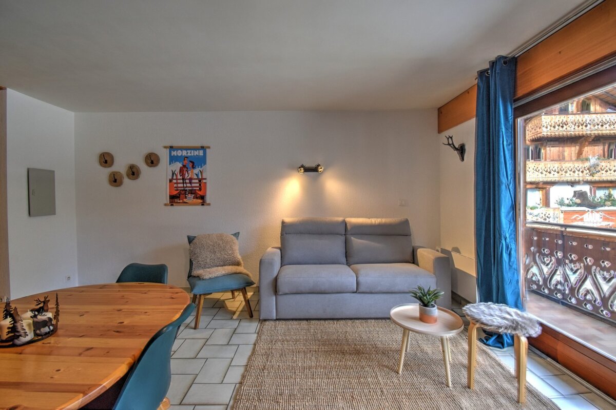 A bright living room features a dining area, grey sofa, Morzine ski poster, and a balcony offering a view of traditional wooden chalets.