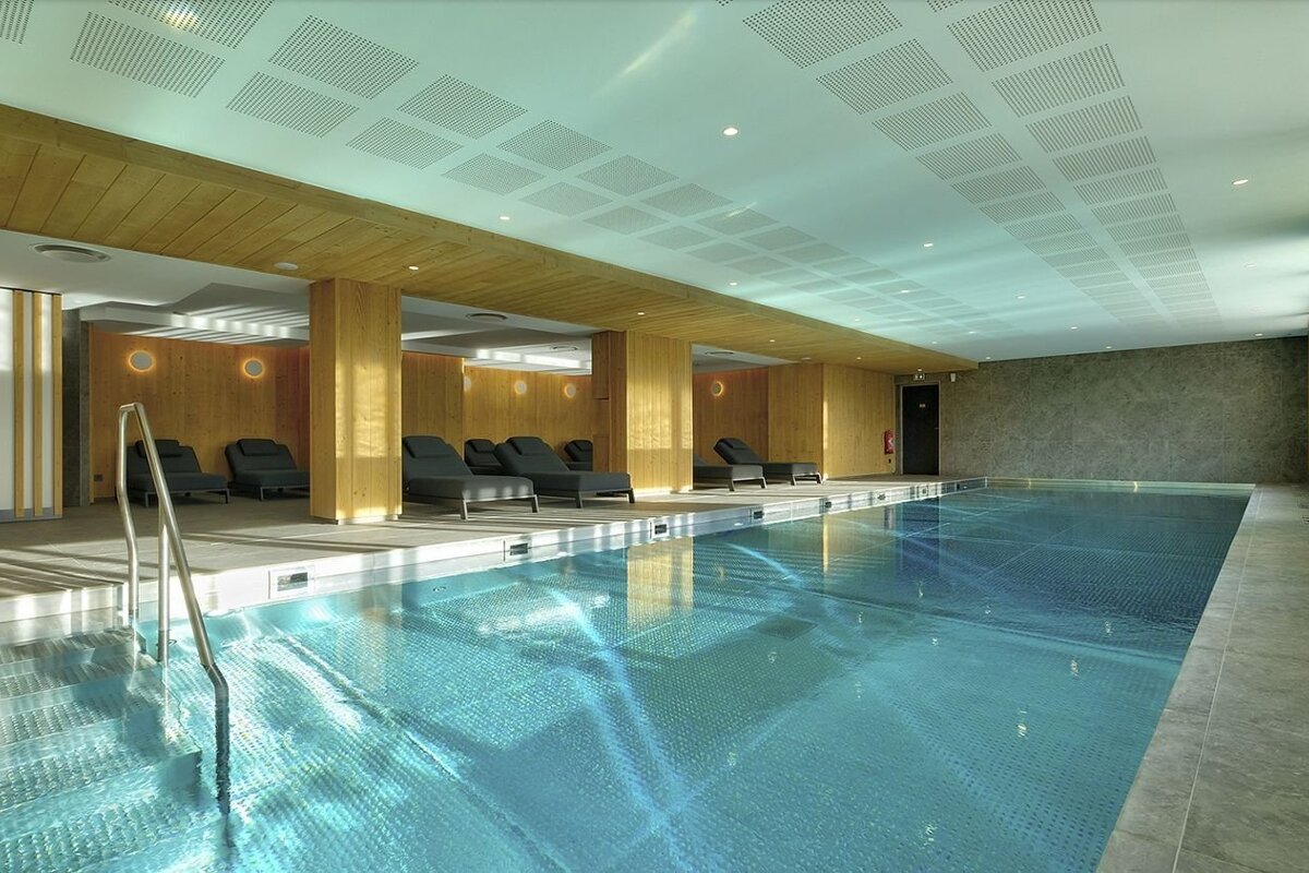 A modern indoor swimming pool with serene blue water, lounge chairs, warm wooden walls, and a perforated ceiling, creating a relaxing atmosphere.