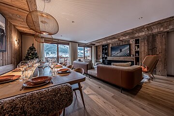 Chic, rustic ski chalet interior. A long dining table is set, leading to a comfy living area with a TV and panoramic snowy mountain views.