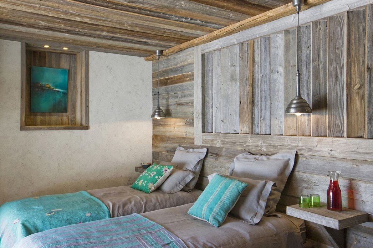 A cozy rustic bedroom with two single beds, featuring reclaimed wood walls, ceiling, and headboards. Teal and green accents, pendant lights, and an abstract painting create a warm ambiance.