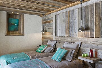 A cozy rustic bedroom with two single beds, featuring reclaimed wood walls, ceiling, and headboards. Teal and green accents, pendant lights, and an abstract painting create a warm ambiance.