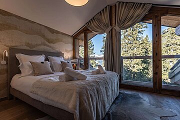 A cozy bedroom with a comfortable bed, warm lighting, and a large window offering a scenic view of snow-capped mountains and pine trees.