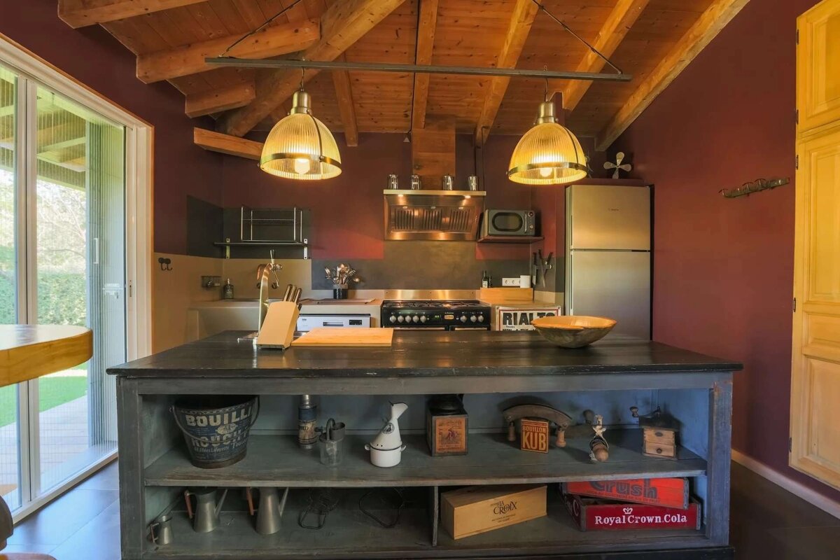 A rustic kitchen with dark red walls, wooden beams, stainless steel appliances, and a large island. Two industrial pendant lights illuminate the space.