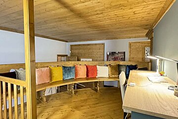 A rustic-modern room with wood-paneled ceiling and floor. A long, curved wooden bench is lined with colorful cushions, beside a bright desk setup.