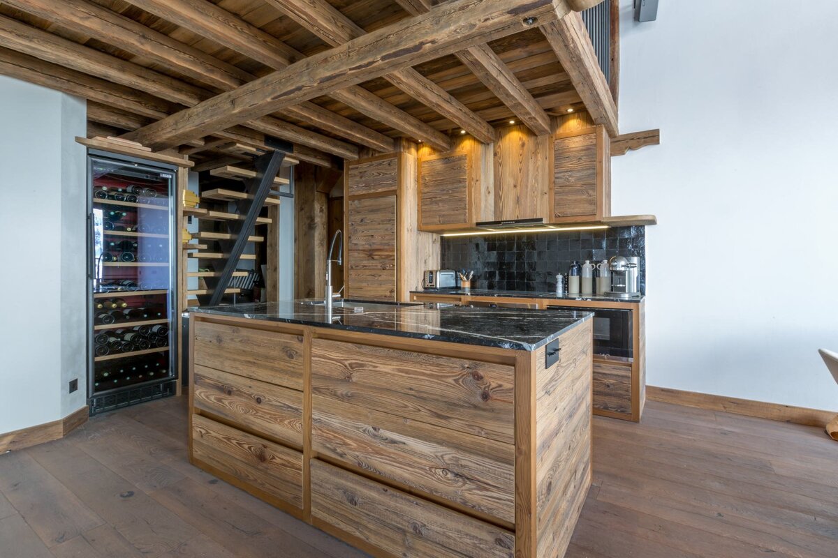 A rustic-modern kitchen featuring natural wood cabinets, a dark marble-topped island, a built-in wine cooler, and exposed ceiling beams.