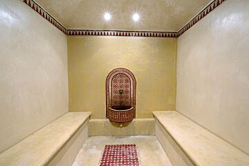 A Moroccan-style hammam featuring a beautifully tiled red and white fountain, built-in benches, and a decorative border against warm yellow and cream walls.