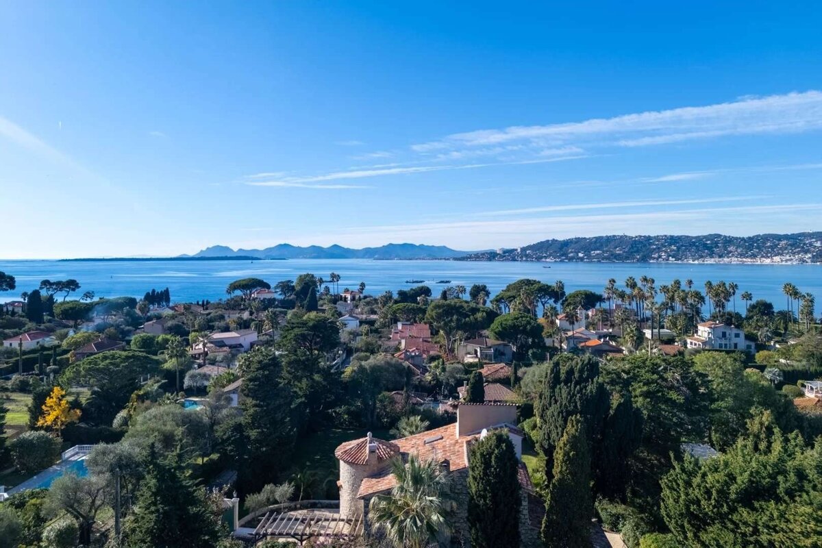 Aerial shot of a vibrant coastal town filled with houses, dense green trees, and palm trees, overlooking a serene blue bay and distant mountains.