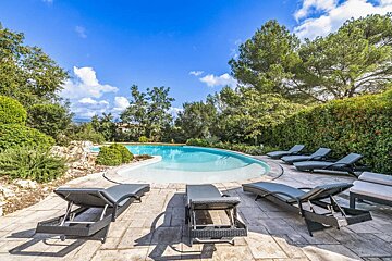A sunny outdoor pool with a white-bottomed pool and dark lounge chairs on a paved patio, surrounded by lush green foliage under a clear blue sky.