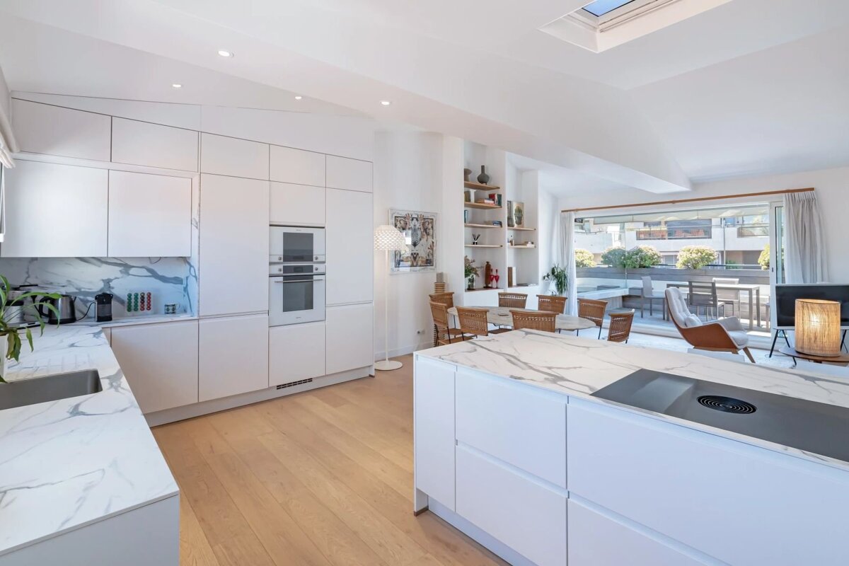 A bright modern kitchen with white cabinets, marble countertops, wooden floors, and an open layout leading to a dining area.