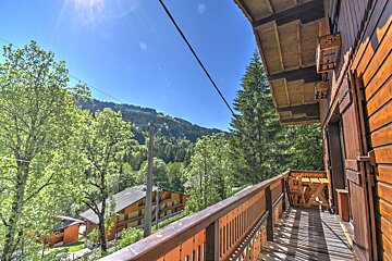 Sunny view from a wooden balcony overlooking lush green mountains, trees, and buildings below, all under a clear blue sky.