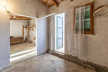 An empty, rustic room with concrete floors, exposed wooden ceiling beams, and unfinished white walls. A window with sheer curtains, a doorway.