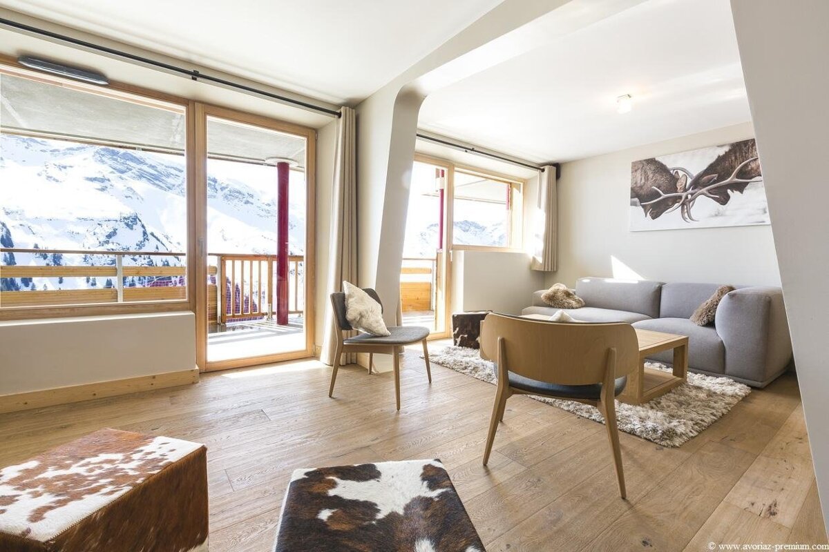 Modern living room with a grey sofa, light wood floors, and cowhide ottomans. Balconies through large windows overlook snowy mountains.
