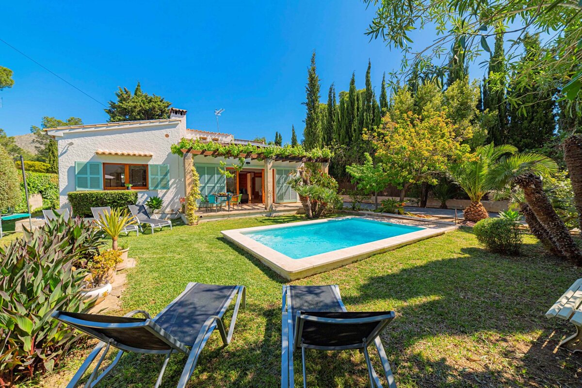 An idyllic white villa with turquoise shutters, a lush green garden, sparkling pool, and sun loungers under a bright blue sky, surrounded by various trees.