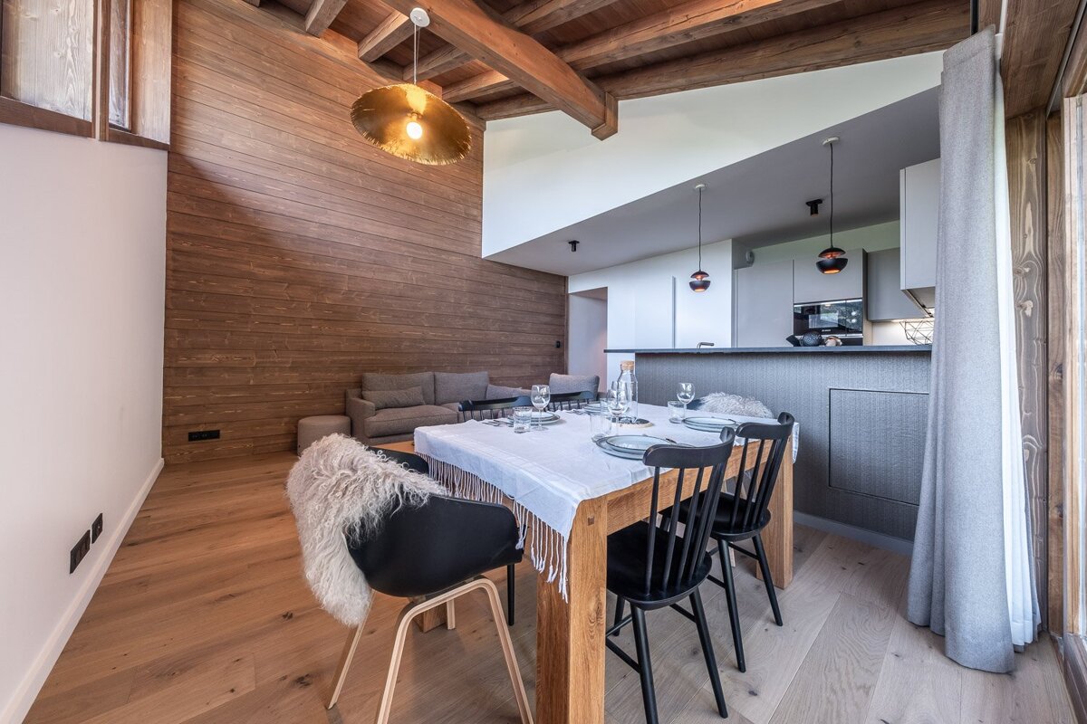 A modern open-plan living space with a wood-paneled accent wall, exposed ceiling beams, a dining table, and a gray sofa. Chic and warm interior.