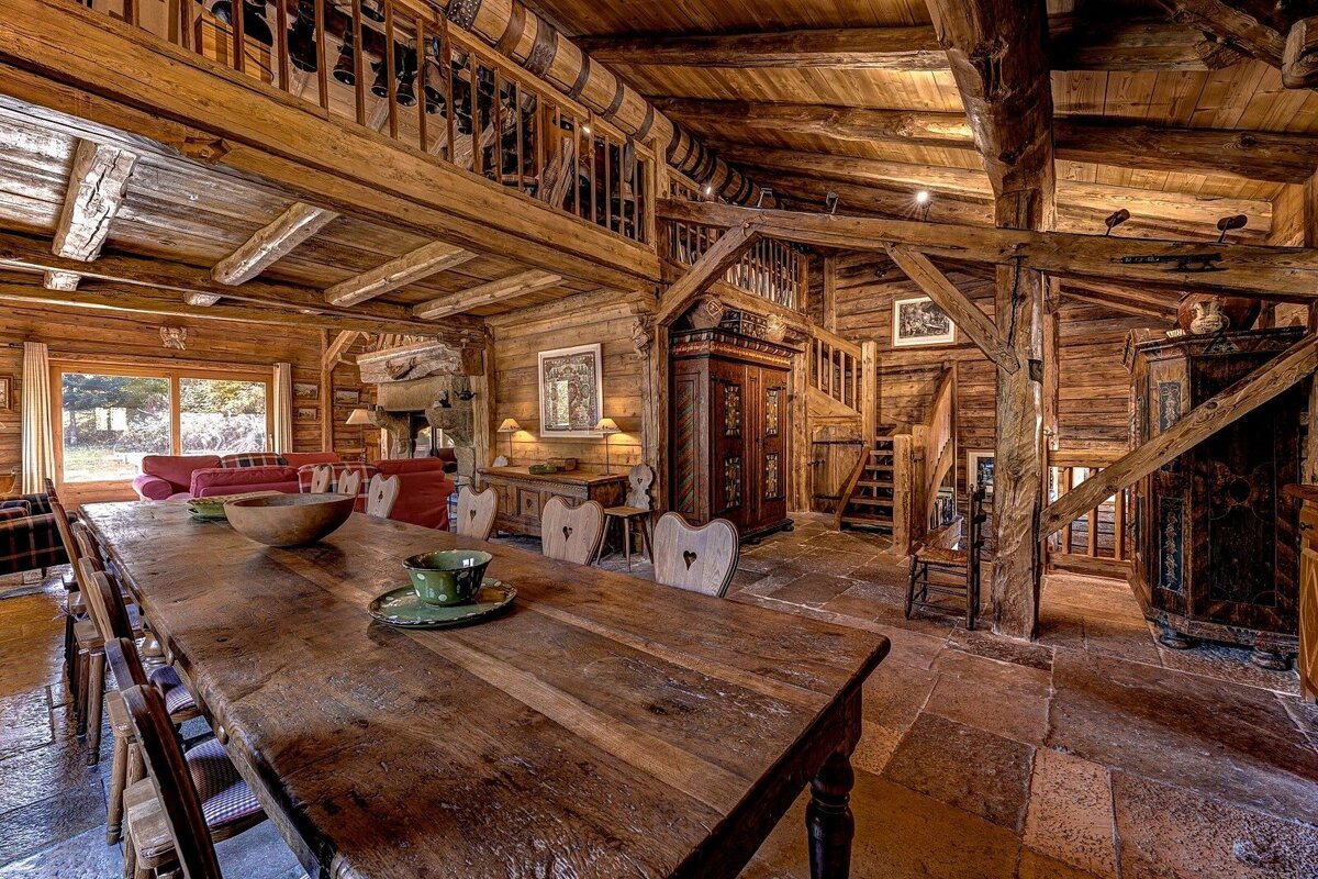 A spacious, rustic wooden lodge interior featuring a long dining table, exposed beams, a mezzanine level, and a warm, inviting ambiance.