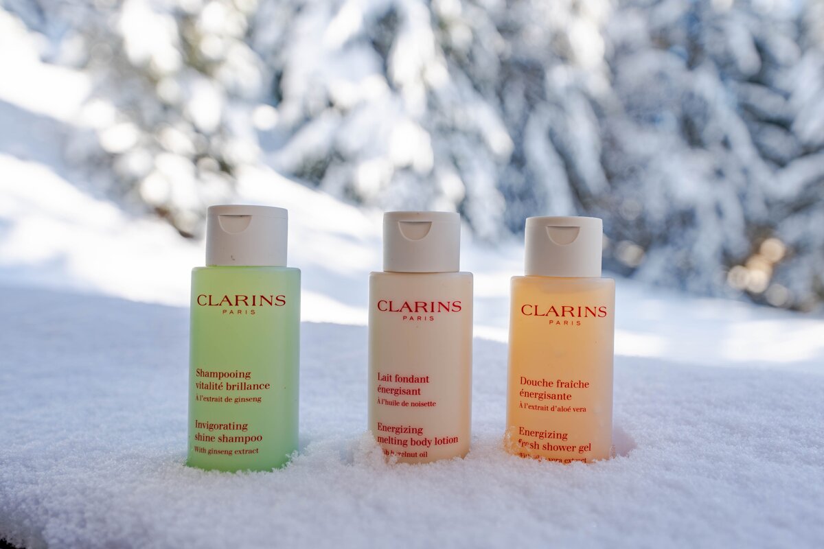 Three Clarins shampoo, lotion, and shower gel bottles stand in fresh snow with a blurred snowy forest backdrop.
