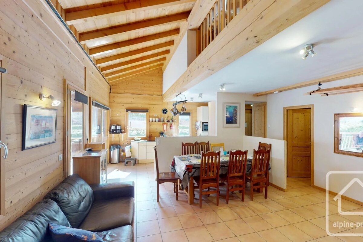 Bright chalet interior with wood-paneled walls, exposed ceiling beams, a leather sofa, dining area, and tiled flooring.
