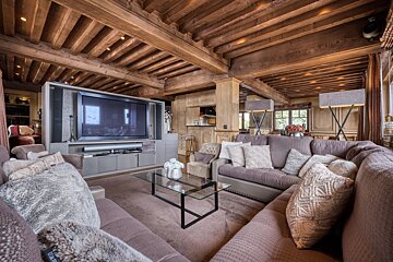 Rustic chalet living room featuring exposed wood beams, a massive TV, large L-shaped sofa, glass coffee table, and a kitchenette view.