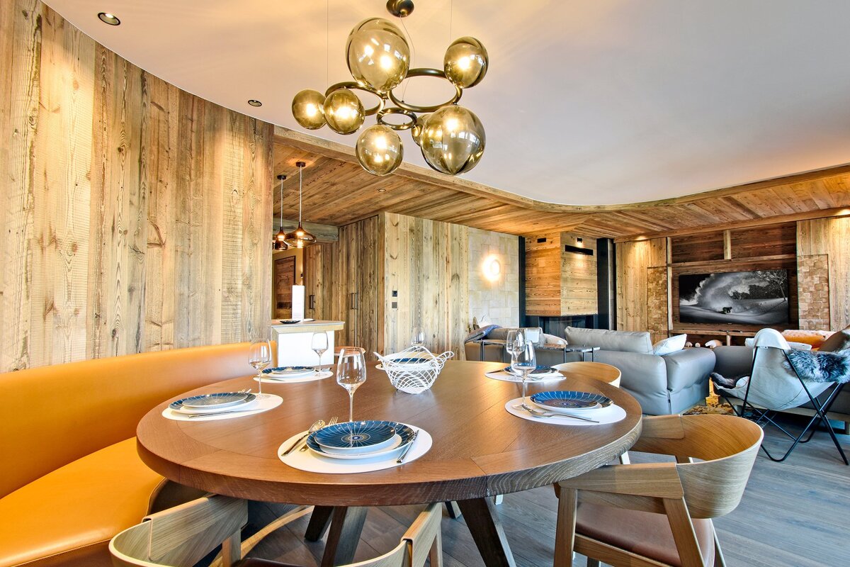 A modern wooden chalet dining room with a round table, orange banquette, and unique chandelier, flowing into a cozy living area.