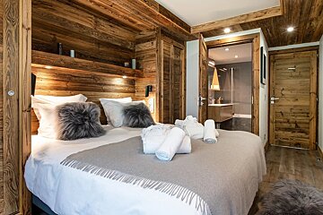 A cozy, modern chalet bedroom with extensive natural wood paneling. A large bed with white linens, grey blanket, and faux fur pillows leads to a visible bathroom.