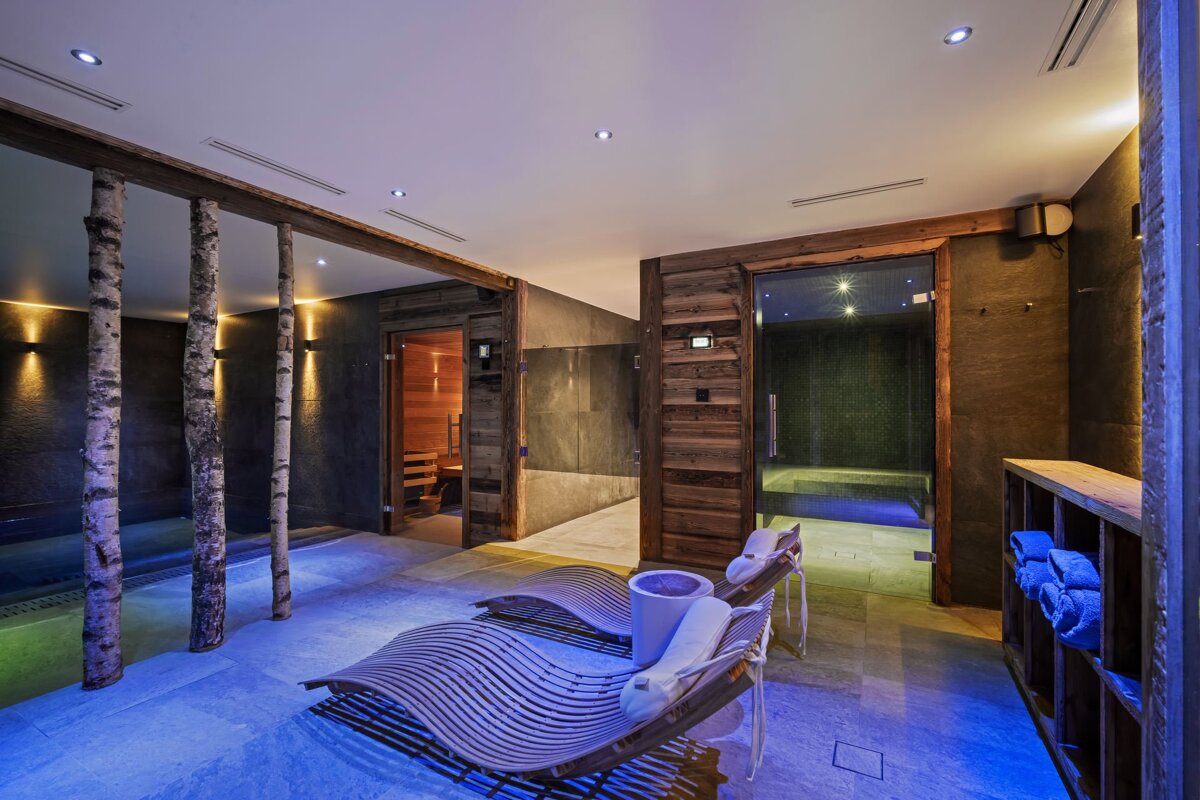 A serene spa area with natural wood, birch pillars, and blue-lit flooring. It features modern loungers, a sauna, and a glass-enclosed steam room.