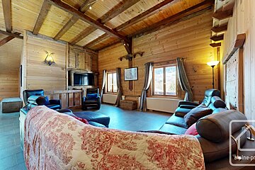 Rustic wood-paneled chalet living room with exposed beams, leather sofas, a TV area, and bright windows overlooking a scenic view.