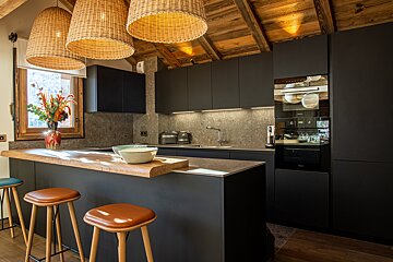 A modern kitchen blends rustic charm with black cabinets, stone backsplash, and wooden ceiling. Wicker lights illuminate a large island with bar stools.