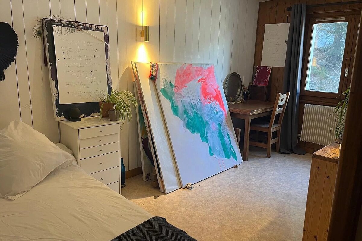 A creative bedroom with a bed, white dresser, large abstract canvases, a wooden desk, and a window. Features plank and wood walls.