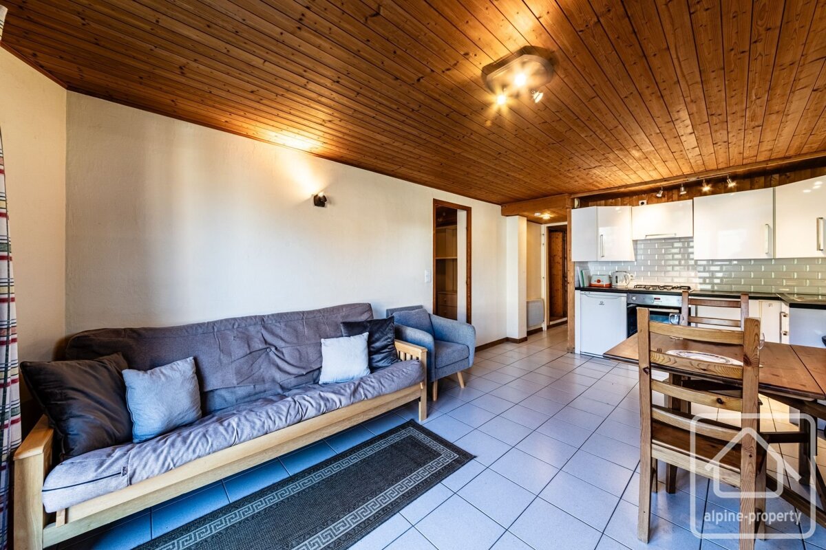 A cozy, open-plan living space features a wooden slatted ceiling, white walls, a grey sofa, tiled floor, and an integrated kitchenette with a dining table.