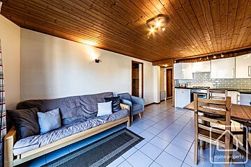 A cozy, open-plan living space features a wooden slatted ceiling, white walls, a grey sofa, tiled floor, and an integrated kitchenette with a dining table.