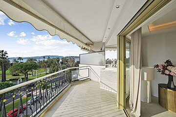 Spacious balcony with a sea view overlooking a palm-lined road, featuring a retractable awning and sliding glass doors to an indoor area.