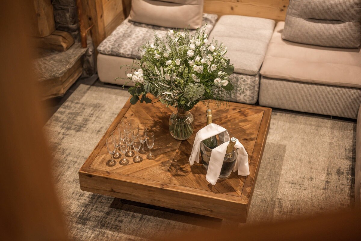A wooden coffee table featuring a large bouquet of white flowers, champagne in ice buckets, and glasses in a cozy living room.