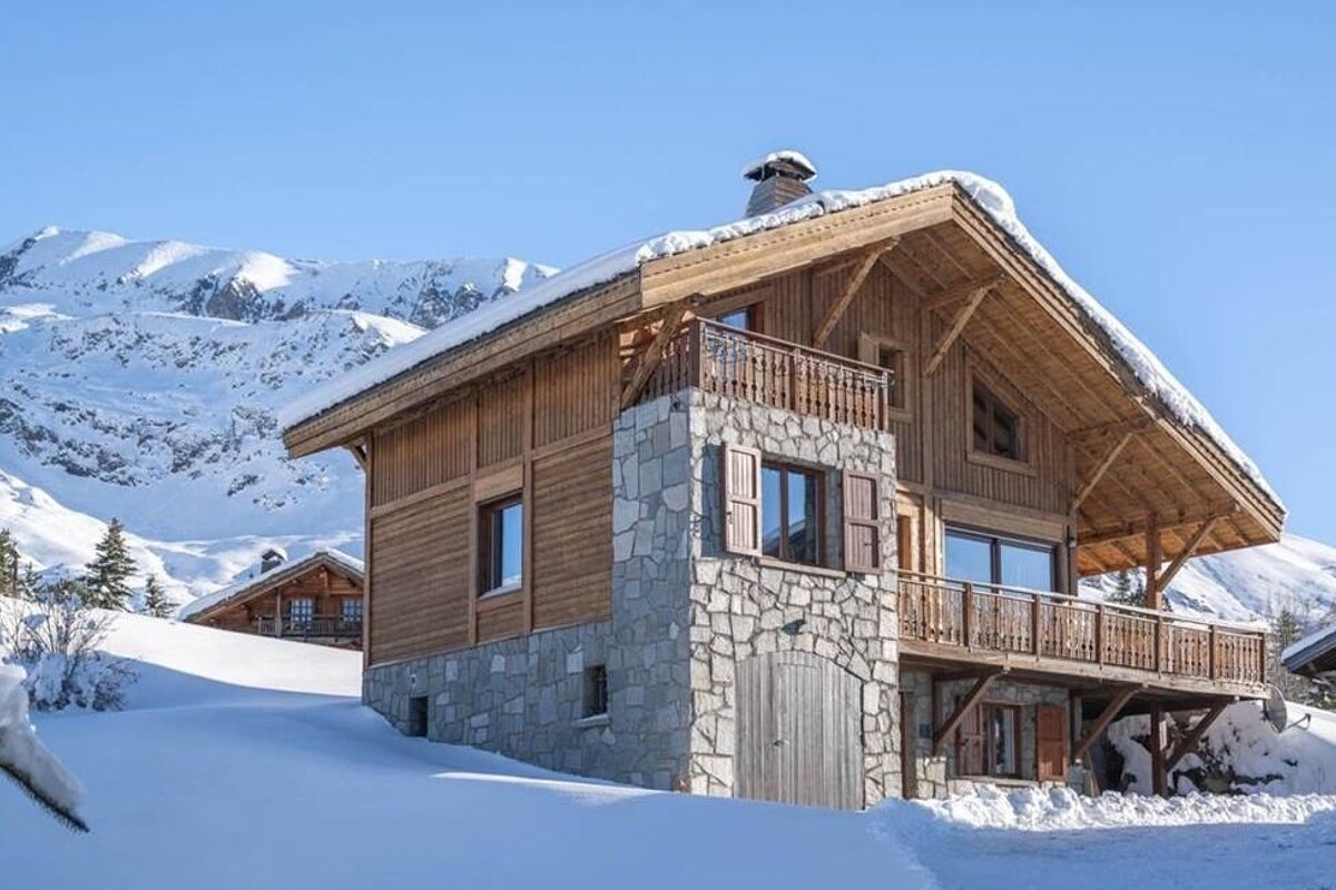 A cozy wooden and stone chalet nestled in a snowy alpine landscape with mountains in the background.
