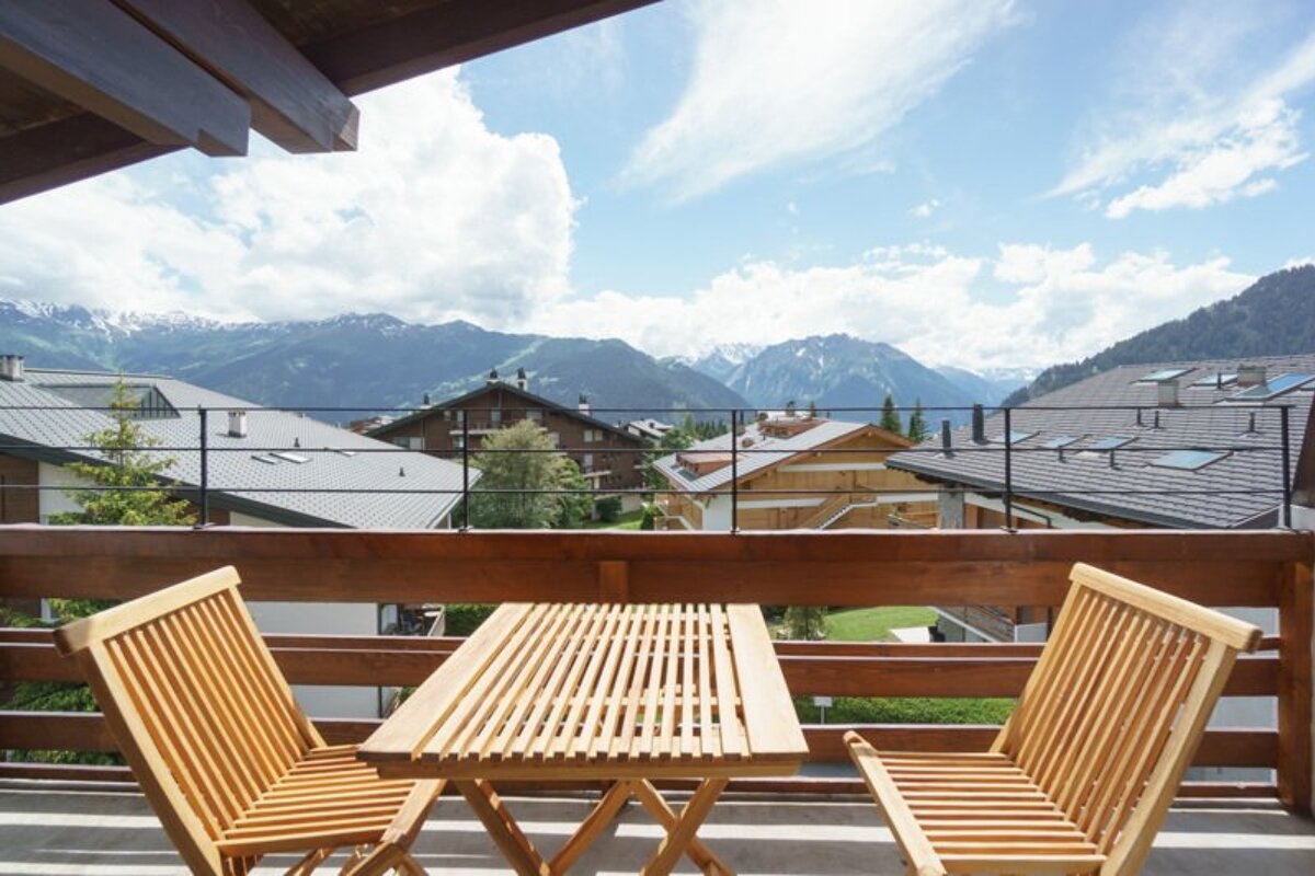 A wooden balcony table and chairs with a scenic view of alpine chalets and mountain peaks under a cloudy blue sky.