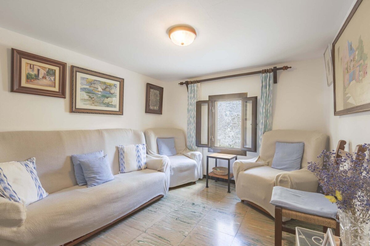 A cozy living room features cream slipcovered furniture with blue pillows, framed artwork, a tiled floor, and an open window with wooden shutters.