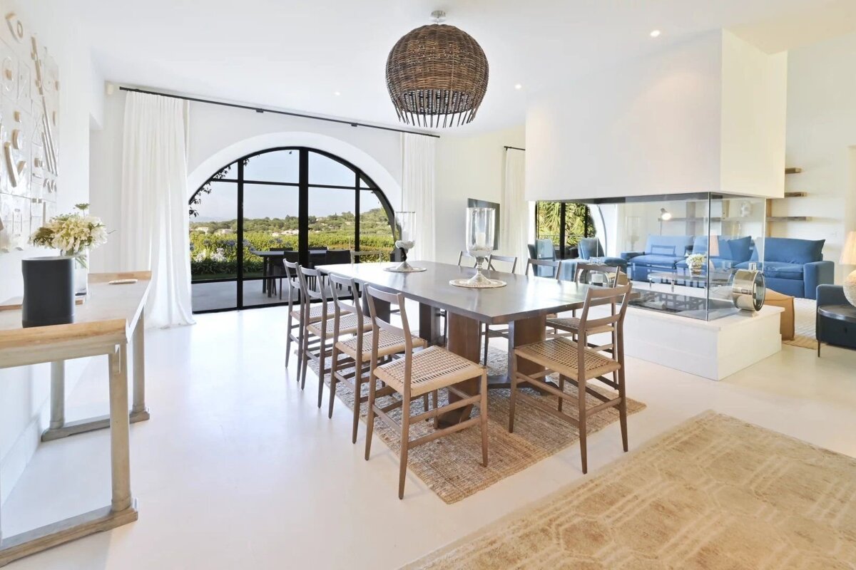 A bright, modern villa dining room featuring a long table, wicker pendant light, and large arched window with scenic views.