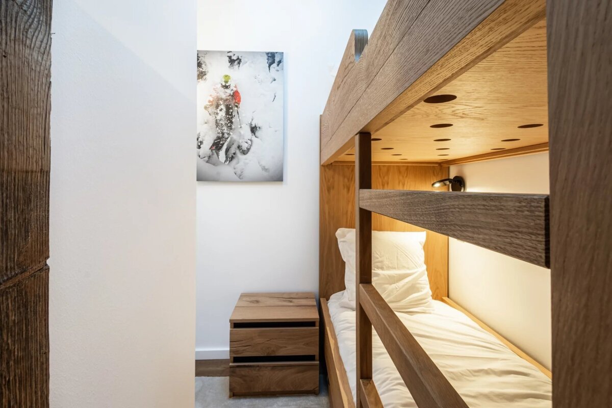 A modern room features light wooden bunk beds with white bedding, a matching nightstand, and a skiing photo on the crisp white wall.