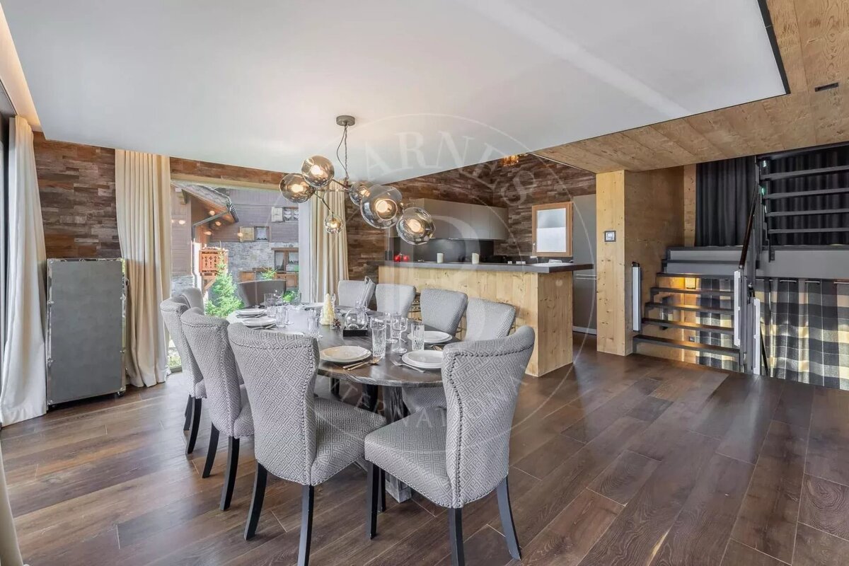 A modern open-plan dining room features a large table, sleek chandelier, kitchen bar, wood-paneled walls, dark floors, and a staircase.