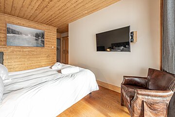 A cozy bedroom featuring a wooden ceiling & wall, a white bed with towels, a wall-mounted TV, and a brown leather armchair. Hardwood floors.
