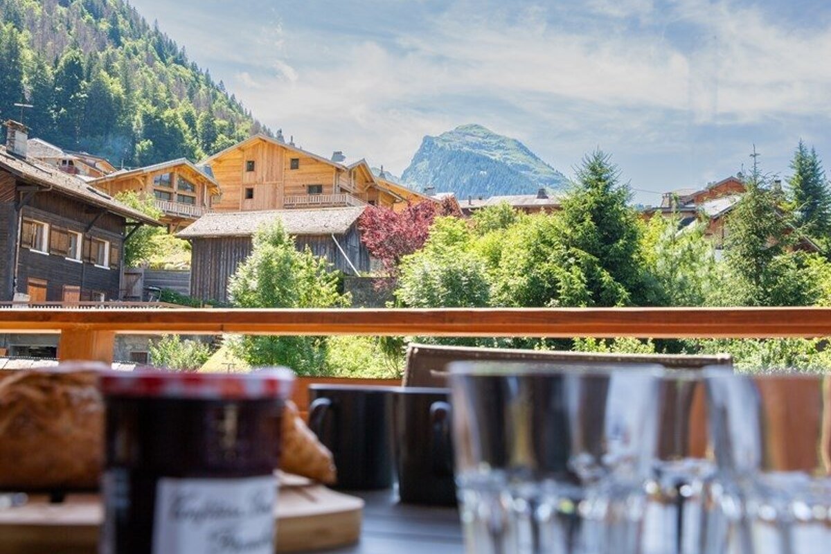 View from a balcony breakfast table of a scenic alpine village with wooden chalets, green trees, and a mountain peak.